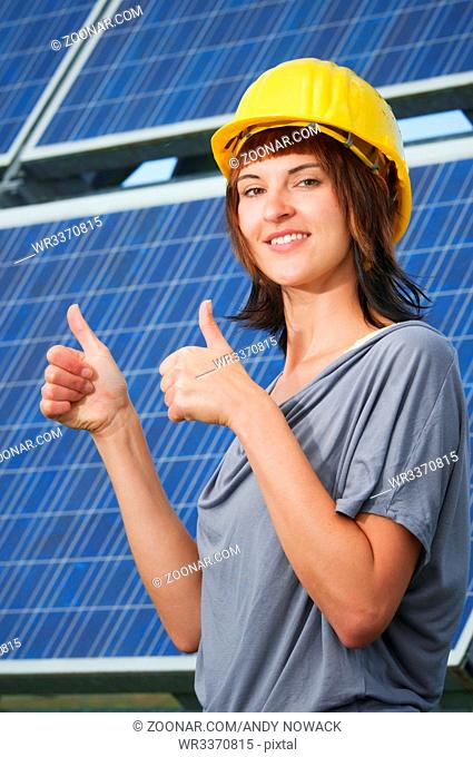 Oberkörper-Ansicht einer jungen Frau mit Baustellenhelm vor einer Photovoltaic-Anlage beide Daumen nach oben zeigend und in die Kamera lächelnd