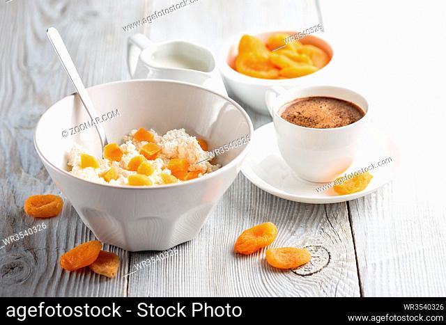 Breakfast from cottage cheese with slices of dried apricot and cup of black coffee on white wooden background
