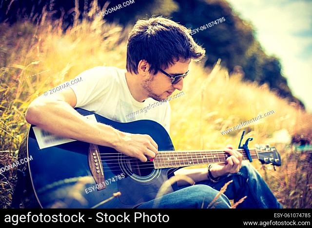 Young man is sitting at a river and playing his western guitar
