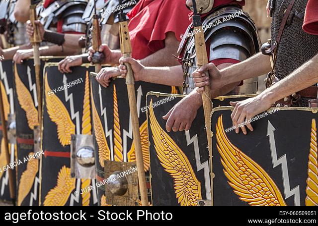 Roman legionaries in formation. Hands holding pila or javelins. Faint eagle-wing and thunderbolt motifs on the scuta