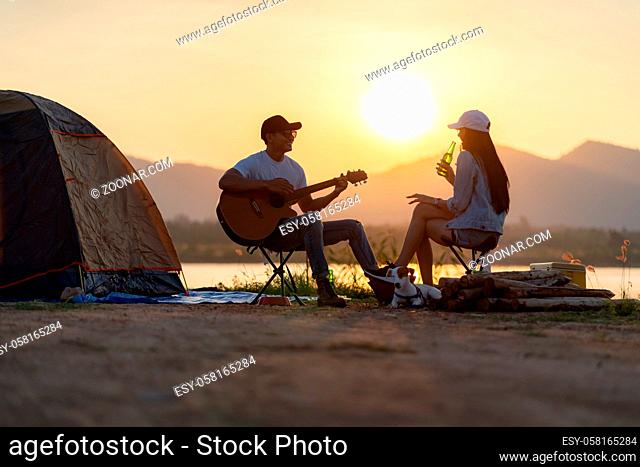 Young adult Asian couple playing guitar and drinking beer beside their tent campsite while sunset. Camping trip with dog outdoor activity campsite concept