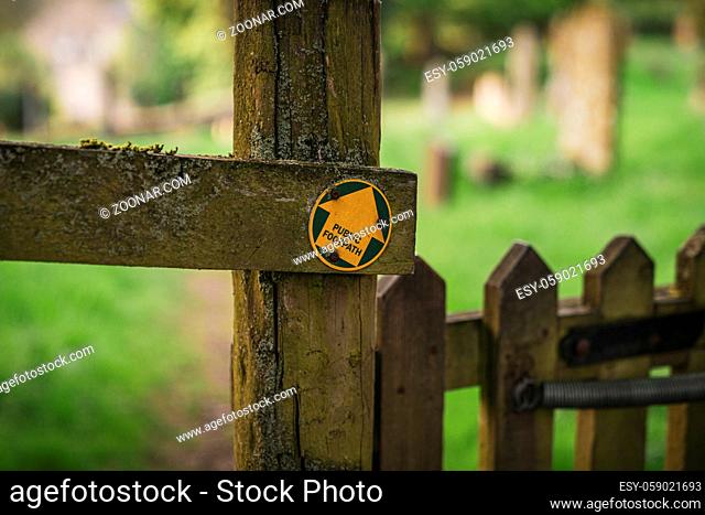 Sign: Public Footpath, seen on a wooden fence, with a blurry cemetery in the background, seen in Aysgarth, North Yorkshire, England, UK