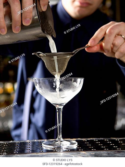 A person pouring a cocktail from a shaker through a strainer into a glass