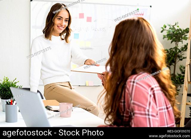 Smiling businesswoman giving notepad to colleague while sitting on tablet at office