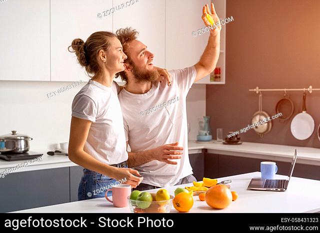 Hiding from a ray of lights shining from a window young couple making breakfast in kitchen at home. Girl hug husband smiling looking away