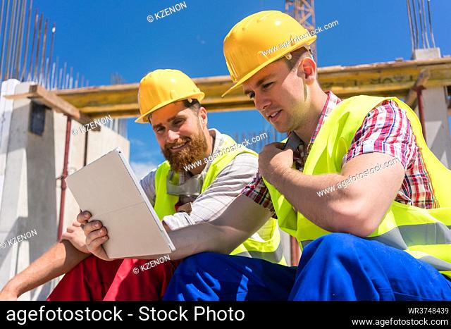 Side view of two workers reading online information or watching a video on a tablet PC, during break at work on the construction site of a contemporary building
