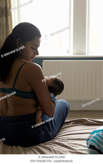 Young mother sitting on the edge of her bed with her baby son sleeping in his arms