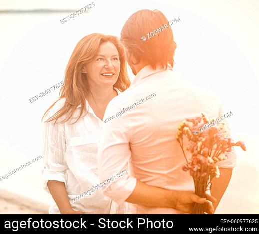 Dating of happy mature couple on seashore. Man about to give bouquet of flowers standing with his back in foreground. Selective focus on smiling mid-aged...
