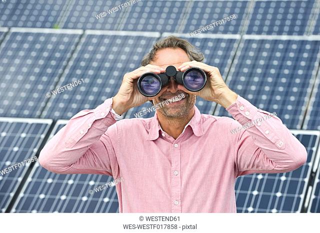 Germany, Munich, Mature man looking through binocular in solar plant, smiling