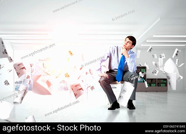 young businessman sits on an office chair. thoughtful and looking up. documents fly around