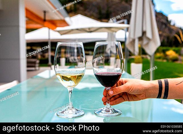 hand holding choosing red wine glass from Two Glasses of Red and white wines on the outdoor restaurant dining table, patio umbrellas in the background