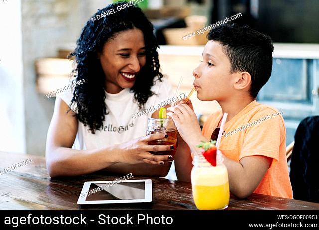 Smiling woman looking while sharing her drinking with son at restaurant