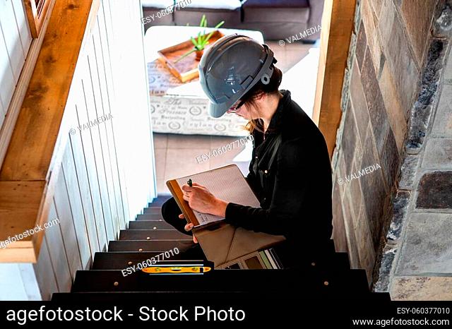 construction inspector woman sitting on the stair and taking notes using clipboard during home pre-purchase inspection, house interior stairs review