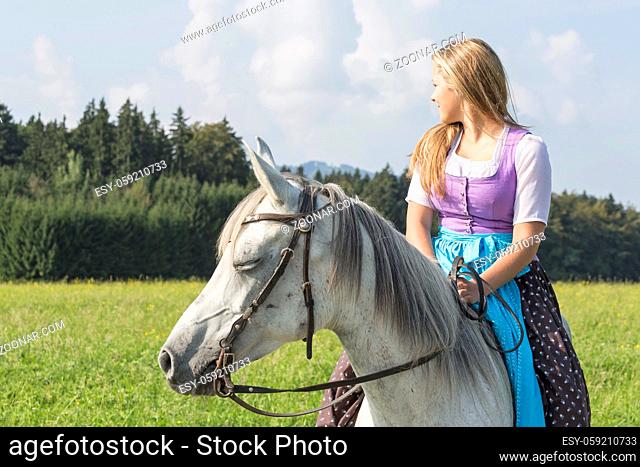 Junge hübsche Frau mit langen blonden Haaren sitzt im Dirndlkleid auf ihrem Pferd