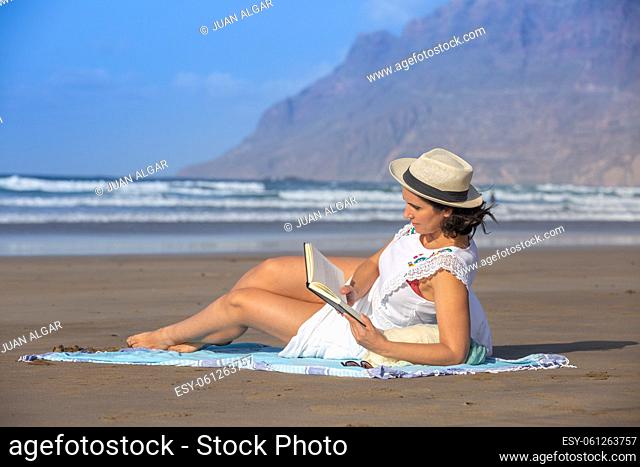 Barefoot middle aged brunette in white dress reading interesting story in book while lounging on sandy beach near waving sea on sunny day on resort