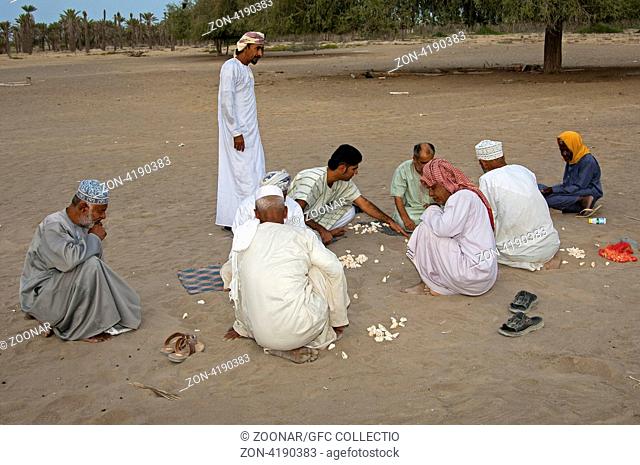 Eine Gruppe omanische Männer spielt Hawalis mit Muscheln im Sand, omanische Variante des Mancala-Spiels, Sultanat Oman / A group of Omani men playing Hawalis...