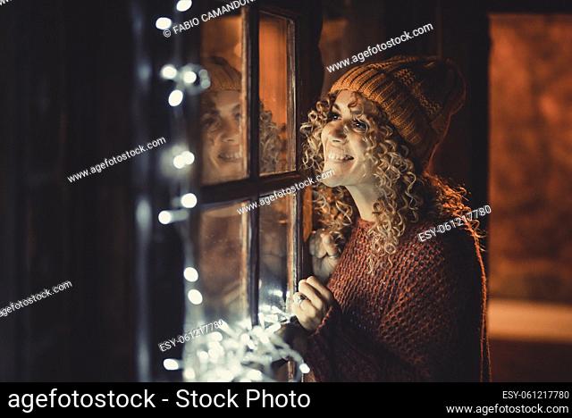 Cheerful adult woman look outside the window at home waiting and smiling for friends to celebrate. Winter leisure indoor lifestyle for christmas or new year eve...