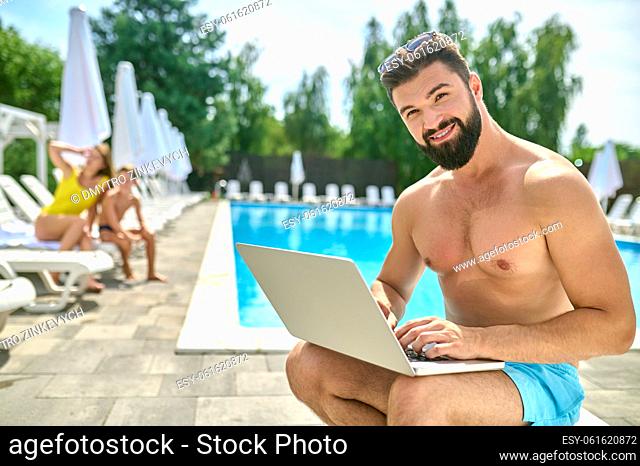Smiling pleased male vacationer with the laptop sitting in the deckchair by the swimming pool and looking ahead