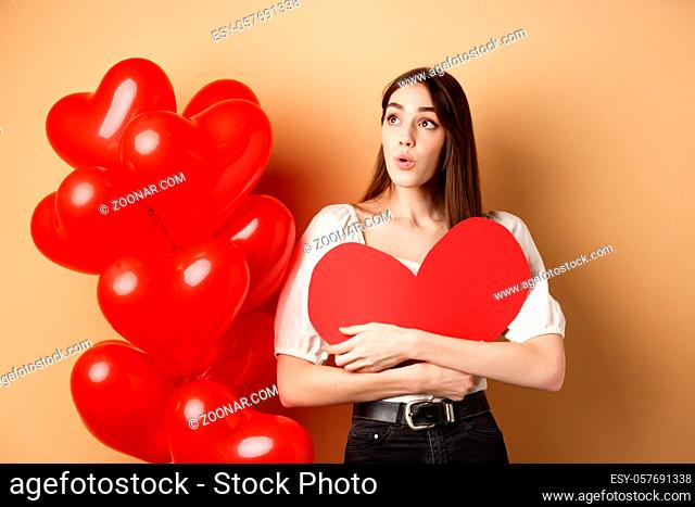 Valentines day and love concept. Intrigued tender girl hugging big red heart cutout and stand near balloons, looking aside at logo, saying wow, beige background