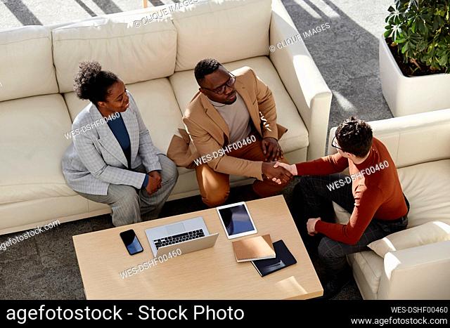 Businessmen shaking hand sitting with colleague on sofa in modern office
