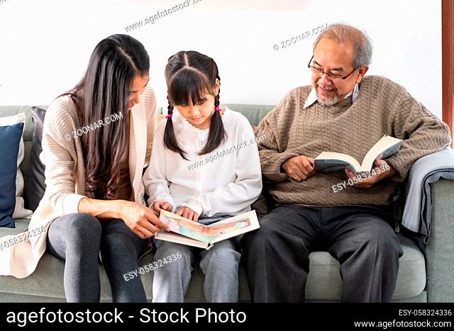 Asian cute girl reading story cartoon book with her mom in living room with grandfather reading fiction book in background