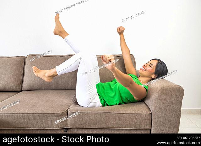 Side view of cheerful barefoot Hispanic female in casual clothes smiling and closing eyes while falling on sofa with raised arms and legs in living room at home