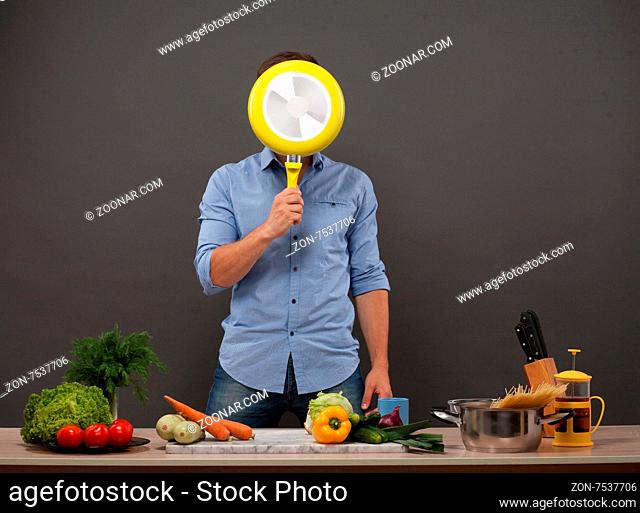 Young chef hiding his face with yellow-coloured pan isolated on dark grey. Man in blue shirt posing in kitchen studio with many products on the counter