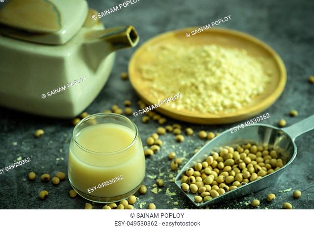 Soymilk in the glass and the kettle is placed beside. Soybean powder is crushed in a wooden dish and has scattered soy beans on the table in morning light