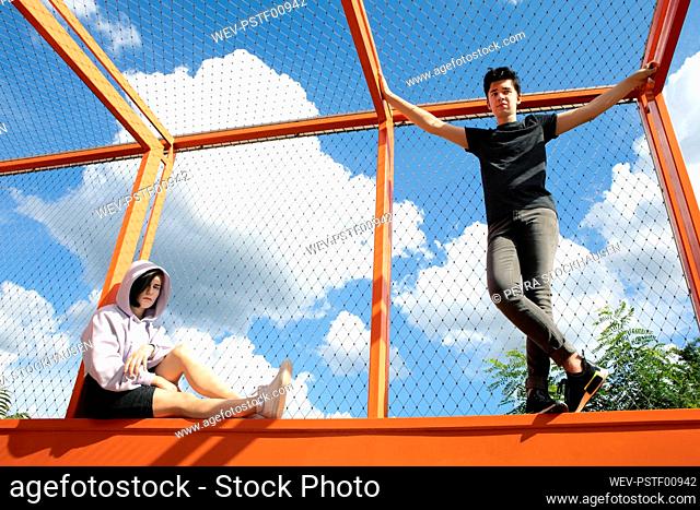Teenage couple by cloud sky on fenced bridge