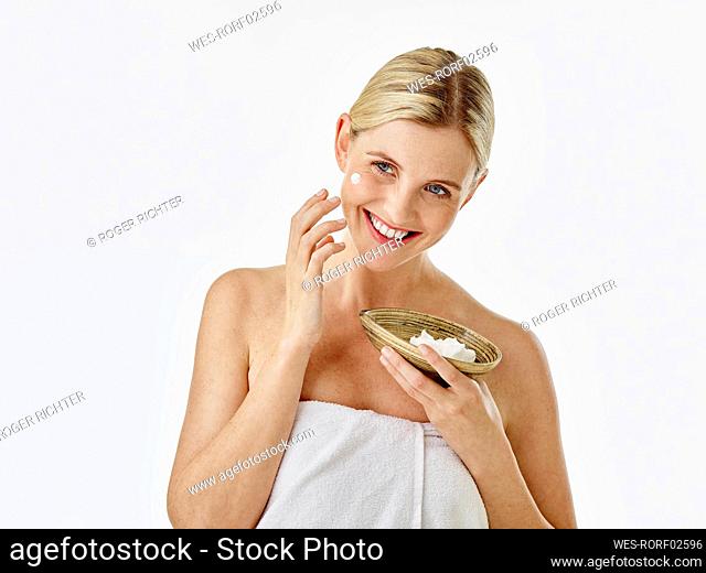 Smiling woman applying face cream while standing against white background