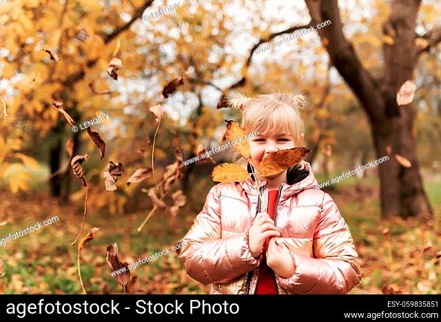Happy little blonde girl plays with yellow autumn leaves in the garden. The child smiles and has fun in park. Playful kid throwing up leaves in autumn season...