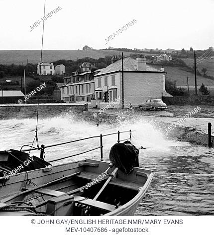 Waves crashing in over a slipway within a small unidentified harbour in Cornwall, with two boats resting in the foreground, and a few houses in the background