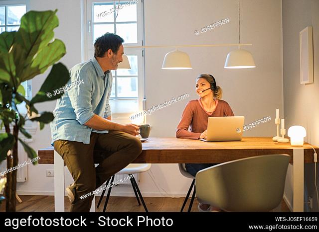 Happy woman with headset talking to man sitting on desk at home office