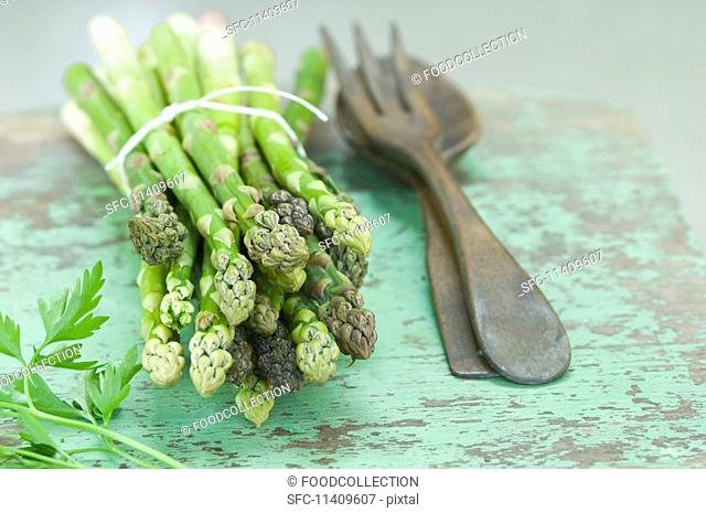 A bunch of green asparagus and parsley on a rustic wooden table with salad servers