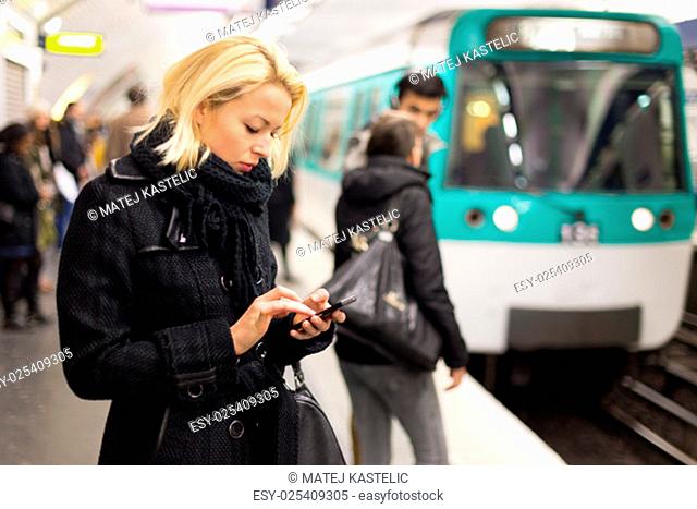 Young woman in winter coat with a cell phone in her hand waiting on the platform of a railway station for train to arrive. Public transport