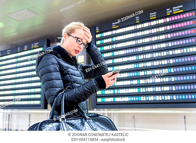 Stressed worried woman in international airport looking at smart phone app information and flight information board, checking her flight detailes