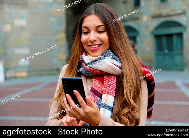 Portrait of beautiful young woman with coat and scarf in city checking her phone