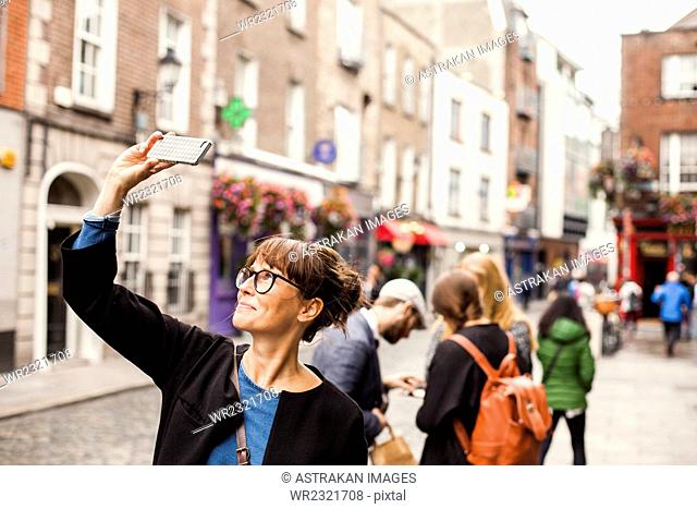 Smiling woman taking selfie with friends standing in background on city street