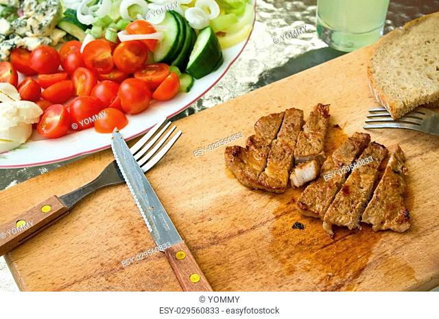 Horizontal photo of pork steak cut in stripes on chopping boar. Knife and fork are next to meat plus bread and several vegetable as tomatoes