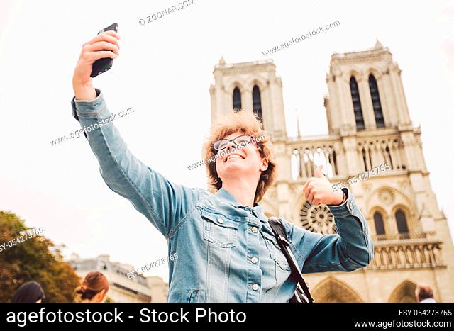 Tourist in Paris making funny selfie near Notre Dame Cathedral. Beautiful young Caucasian tourist woman with backpack in Paris making funny selfie hand holding...