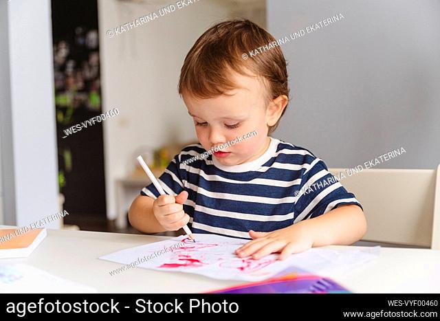 Cute boy drawing on paper with pencil sitting on table at home
