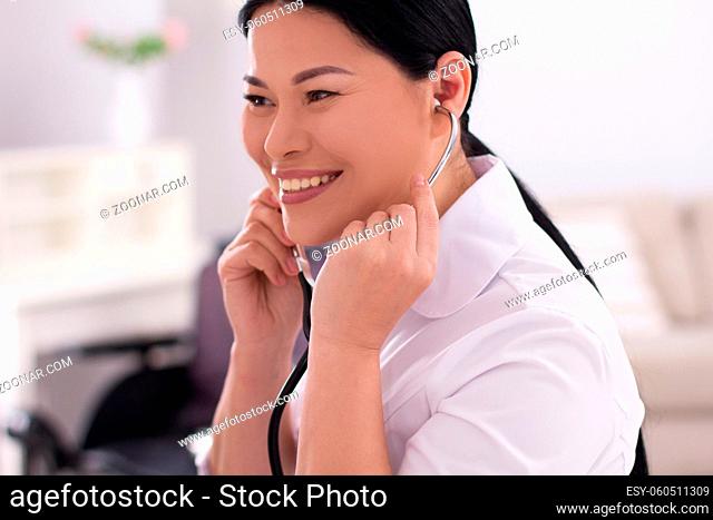 Cheerful asian nurse holding stethoscope. Beautiful asian doctor with her black hair in sleek bun holding stethoscope at her ears smiling wide