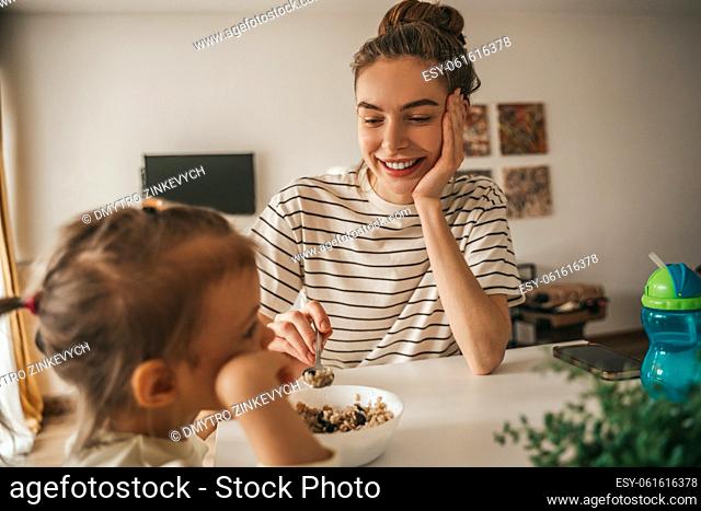 Caring young mother giving a spoon of porridge to her child seated at the kitchen table