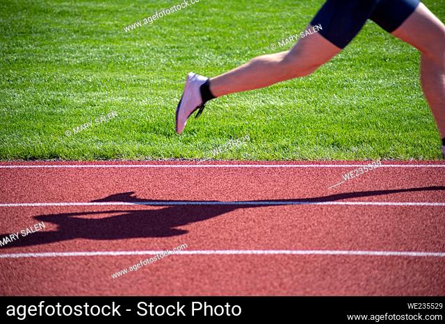 Motion blur on runner's foot and leg as it moves across the image with the runner's shadow on the track. Crisp horizontal lane lines and green grass background...