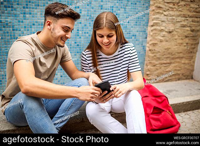 Couple using mobile phone while sitting on footpath