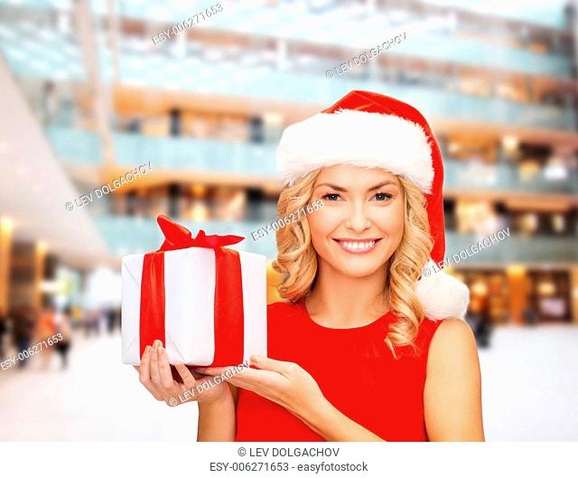 christmas, holidays, celebration and people concept - smiling woman in santa helper hat with gift box over shopping center background