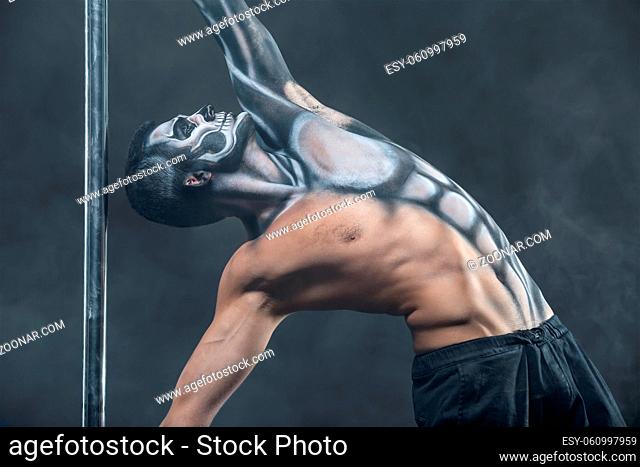 Strong pole dancer with horrific body-art next to a pylon in the dark studio with a smoke on the background. He holds the pylon with the hands and looks up