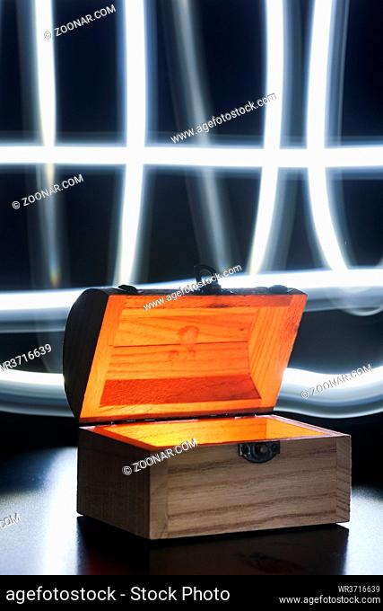 Kleine Holztruhe innen beleuchtet auf blauen Untergrund mit gekreuten Licht-Spuren vor schwarzem Hintergrund. Small wooden chest inside illuminates on a blue...