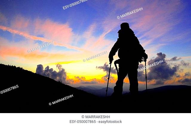 Hiker silhouette with backpack and trekking poles on mountain peak admiring the sunset