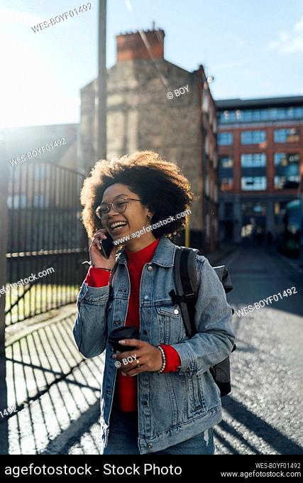 Cheerful afro woman talking over smart phone while standing with coffee cup on street